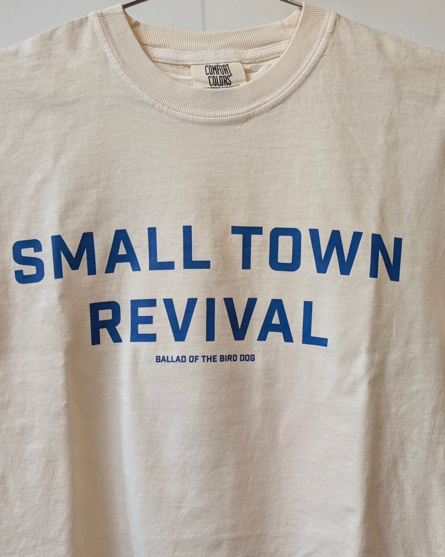 Show your small town spirit with the Ballad of the Bird Dog Shop Shirt—Small Town Revival Crop in beige, featuring blue "SMALL TOWN REVIVAL" text and "BALLAD OF THE BIRD DOG" below.