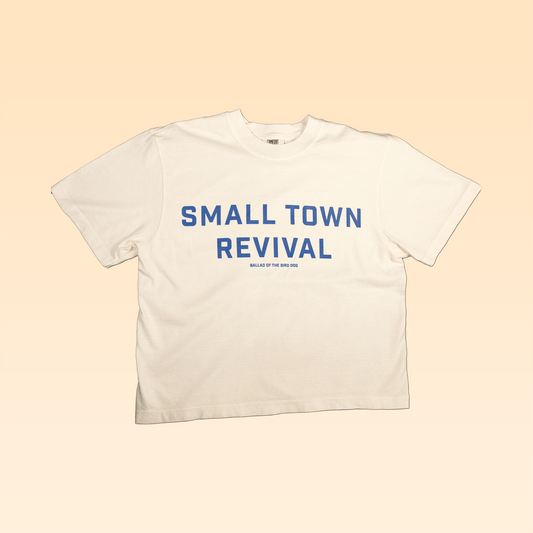 The Ballad of the Bird Dog Shop Shirt | Small Town Revival Crop features bold blue "SMALL TOWN REVIVAL" text on white, laid flat on beige—a perfect crop shirt for showing off your small town pride.