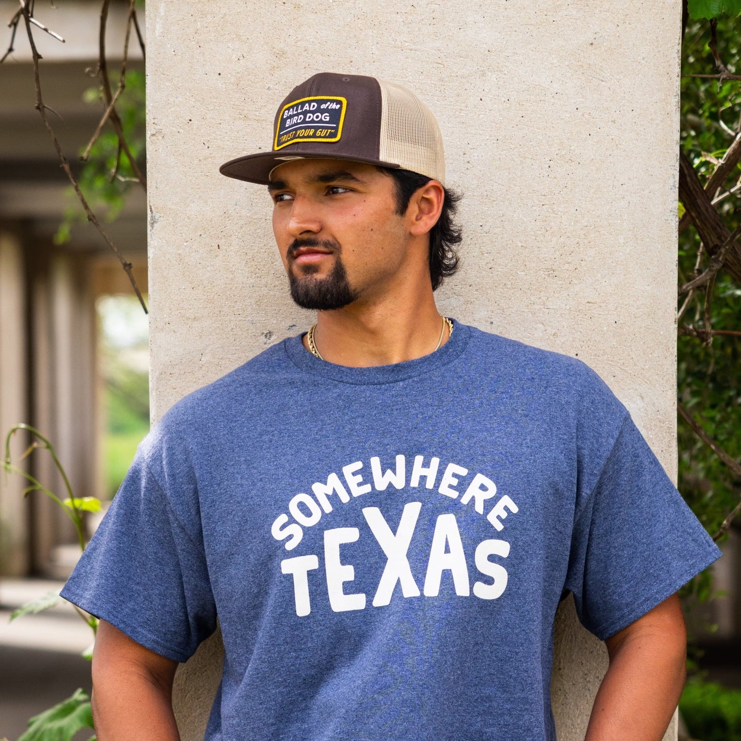 A man in a blue "Somewhere Texas" Shop Shirt by Ballad of the Bird Dog and a trucker hat stands against a concrete pillar with greenery, capturing that relaxed, small-town Texas vibe.