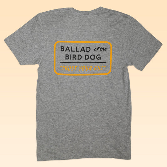 The Shop Shirt | Trust Your Gut Vintage Slate by Ballad of the Bird Dog is a grey tee featuring "BALLAD of the BIRD DOG 'TRUST YOUR GUT'" inside a yellow outlined rectangle on the back—a bold, classic design.