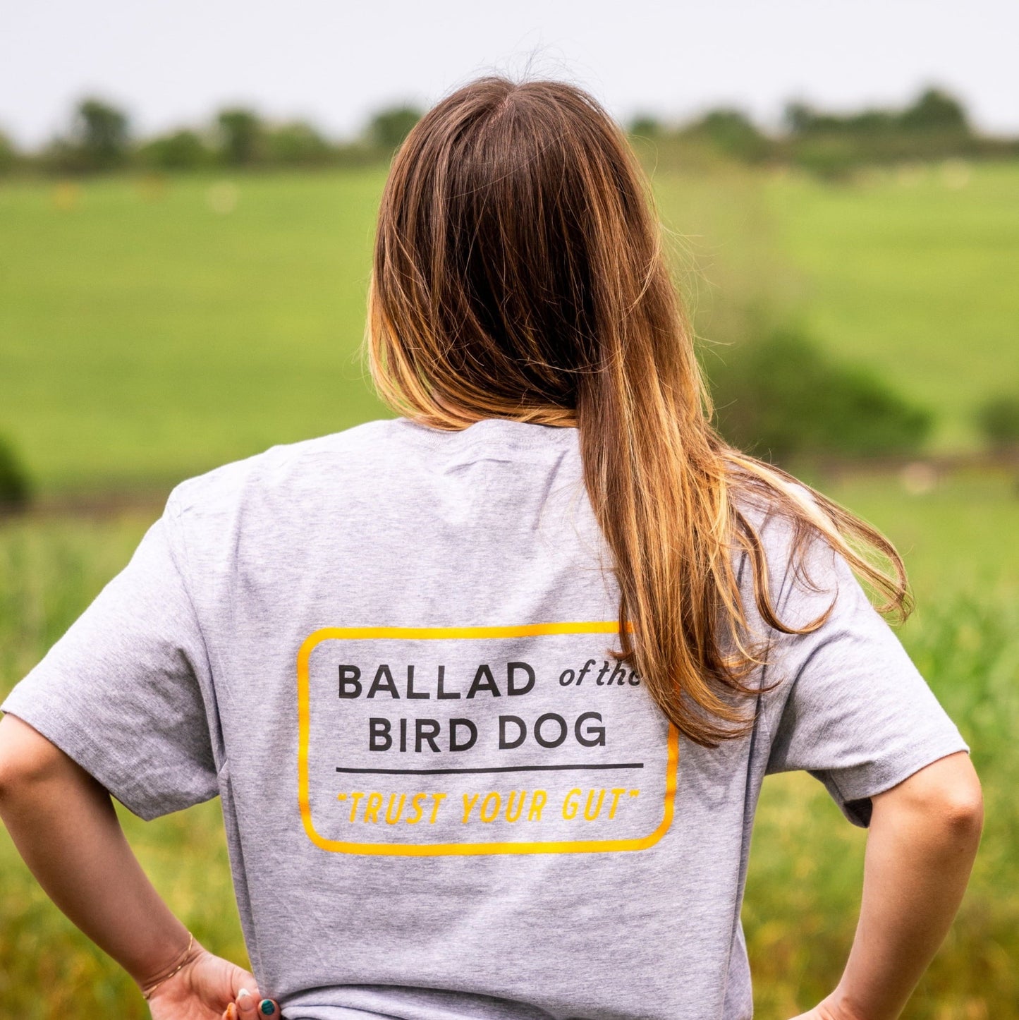 Someone stands outdoors, facing away, wearing the Ballad of the Bird Dog Shop Shirt in Trust Your Gut Vintage Slate with bold black and yellow "BALLAD of the BIRD DOG 'TRUST YOUR GUT'" text on the back.