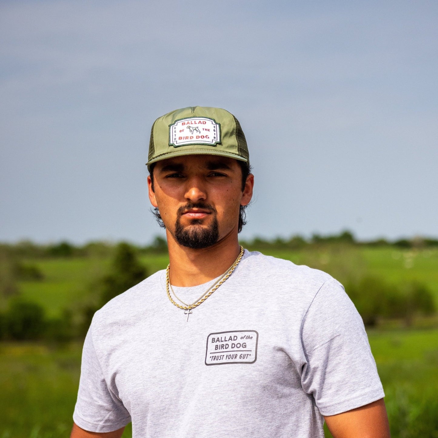 A man with dark hair and a goatee wears a Ballad of the Bird Dog Shop Shirt in Trust Your Gut Vintage Slate and a green cap, standing outdoors in front of a grassy field under a clear sky.