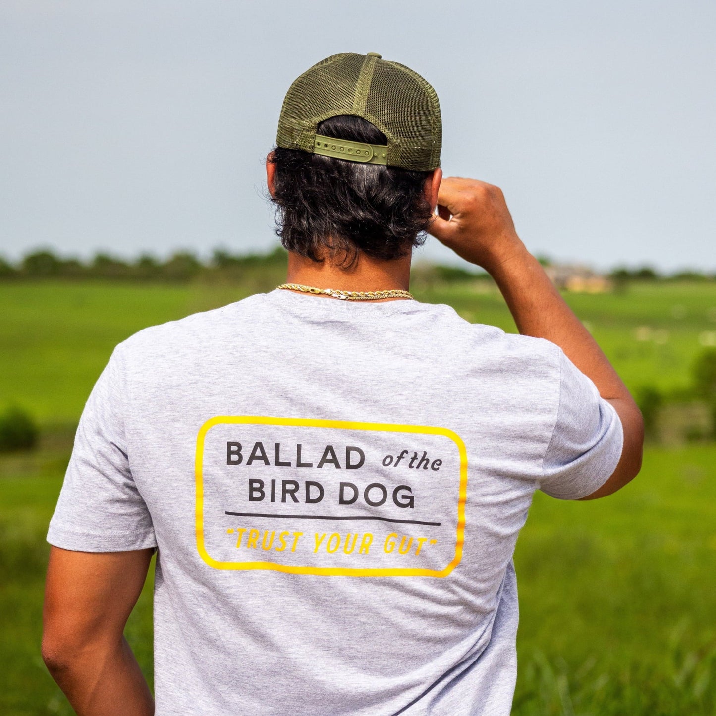 A man wearing the Ballad of the Bird Dog Shop Shirt in Trust Your Gut Vintage Slate and a green cap stands outdoors in a grassy field, facing away from the camera.