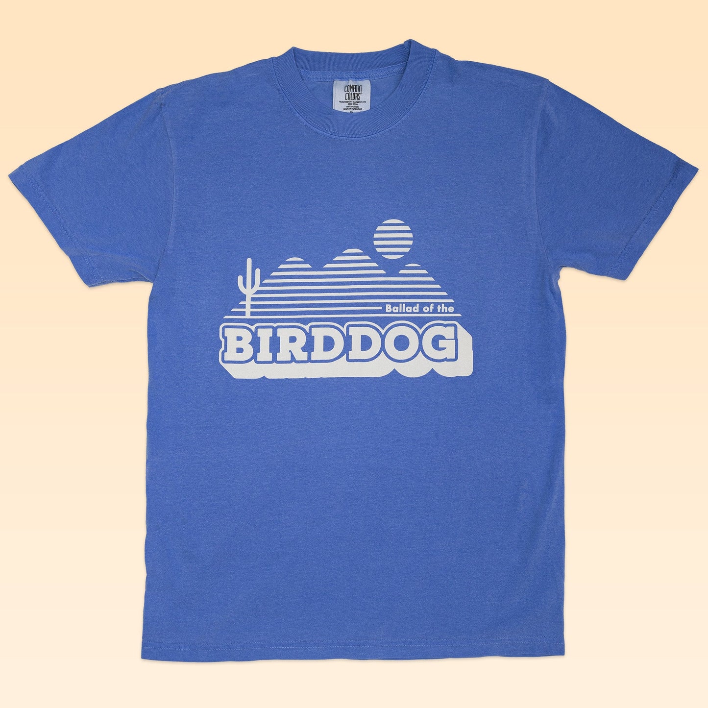 The Shop Shirt by Ballad of the Bird Dog features a blue tee with a white desert scene and "Ballad of the BIRDDOG" graphic, adding vintage southwestern style to your look.