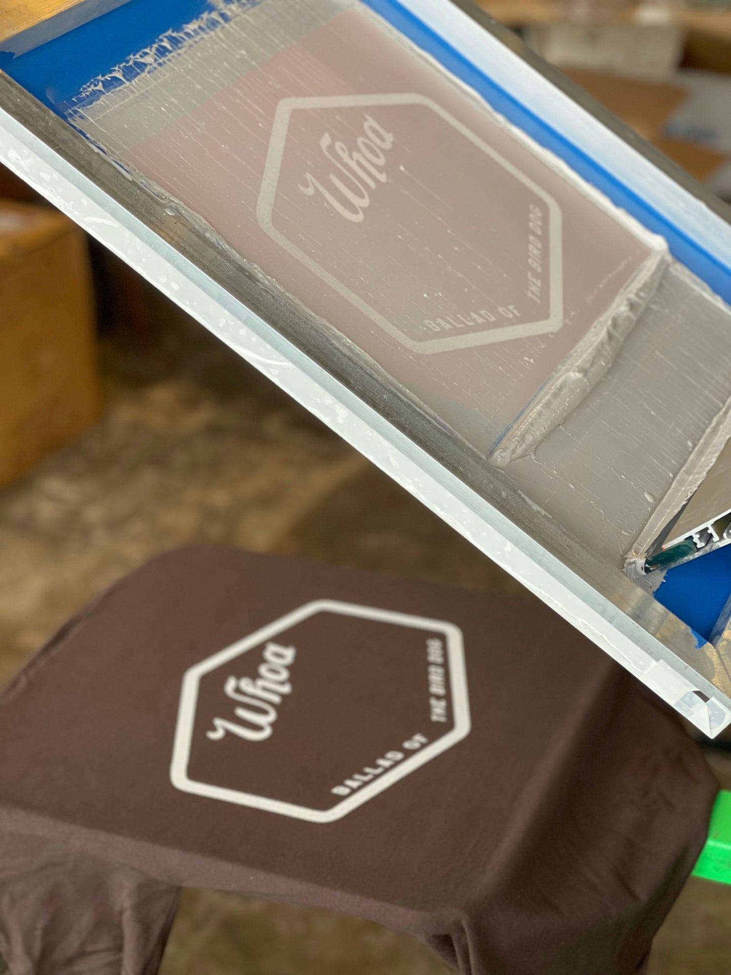 A screen printing frame is set above the Shop Shirt | Whoa from Ballad of the Bird Dog, a brown tee featuring a white "Whoa" logo, ready for printing.