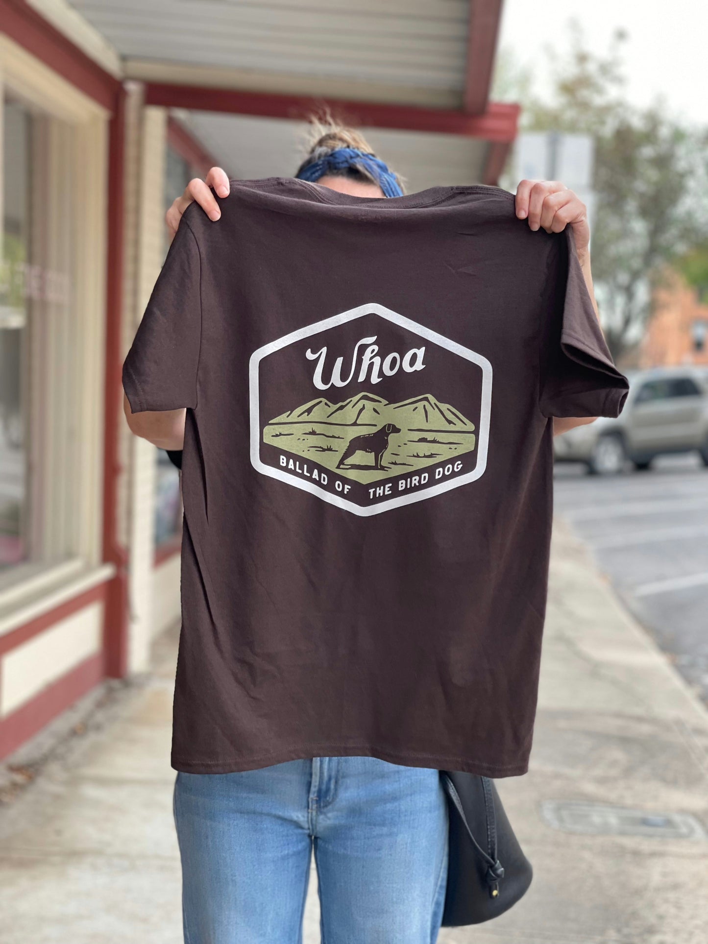 Someone stands on a sidewalk holding up the Ballad of the Bird Dog Shop Shirt | Whoa, which features a graphic of mountains, a dog, and the text "Whoa Ballad of the Bird Dog.