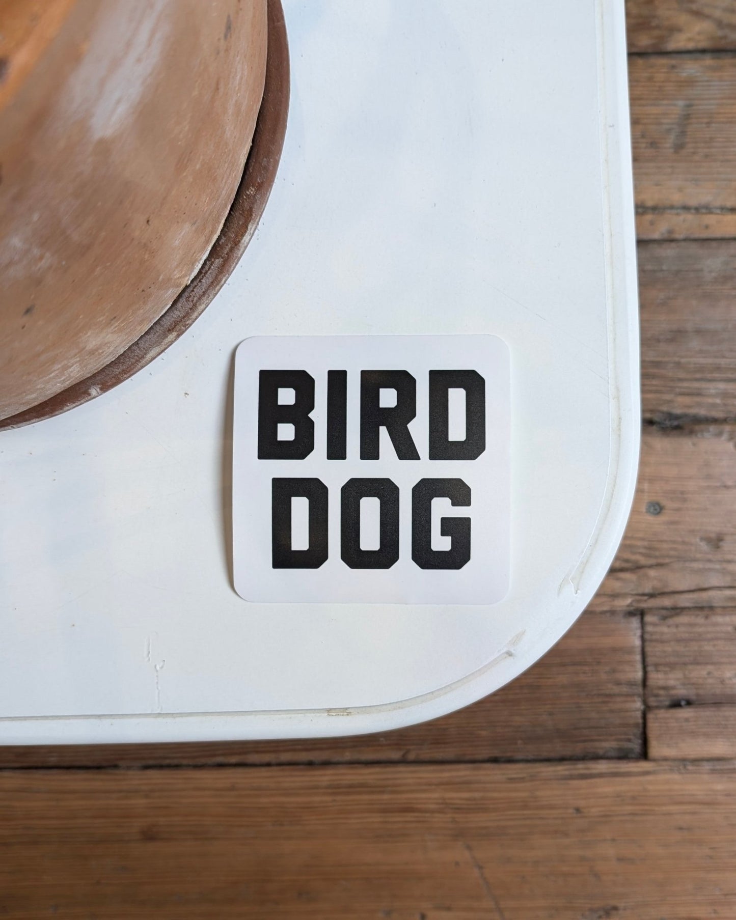 A square white "Shop Sticker | Bird Dog Honor" by Ballad of the Bird Dog with bold black "BIRD DOG" text rests on a white table beside a brown planter, evoking classic bird dog sticker style against wooden floorboards.