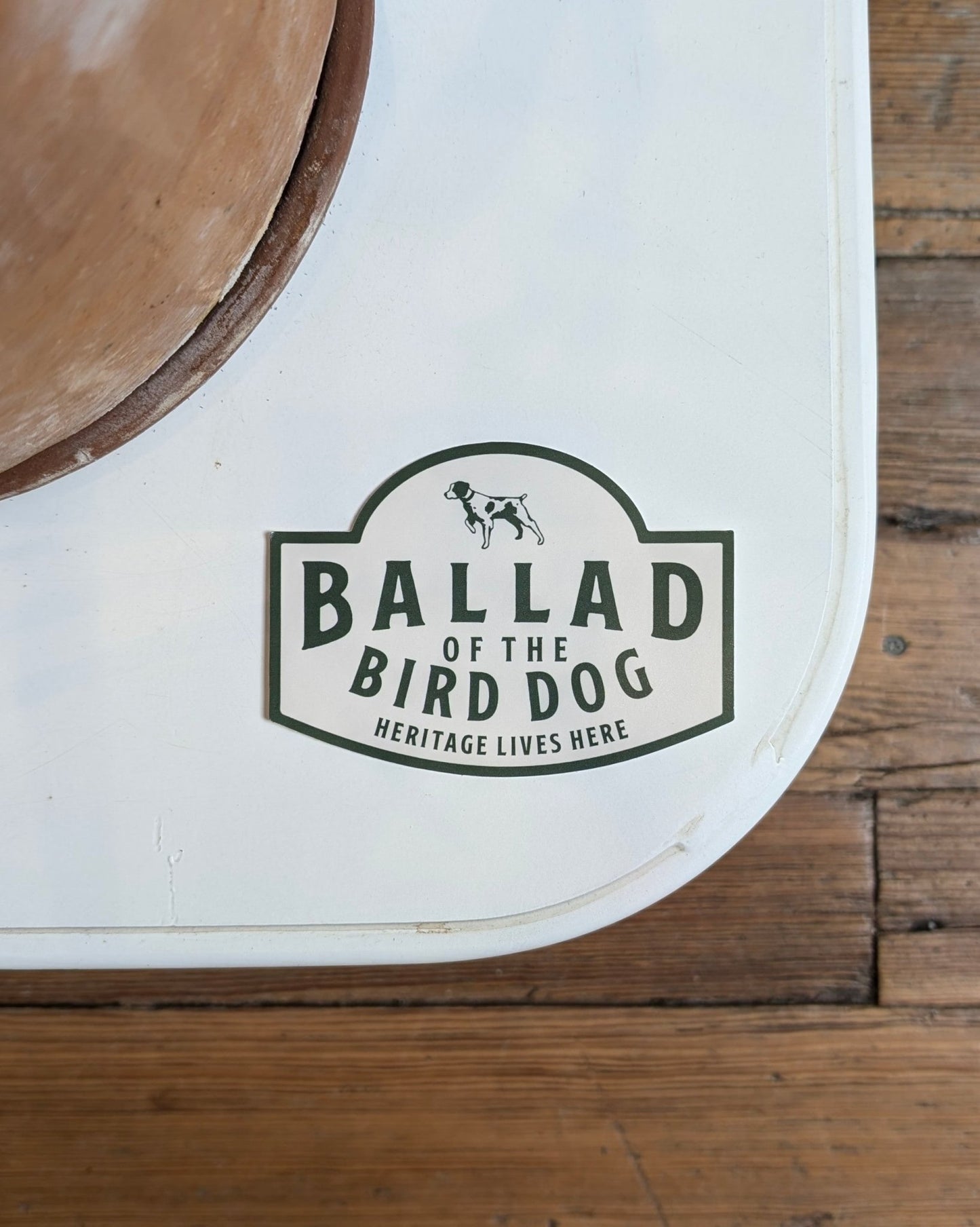 A white surface shows the Ballad of the Bird Dog "Heritage Lives Here" Shop Sticker by a wooden circle on a wood floor.