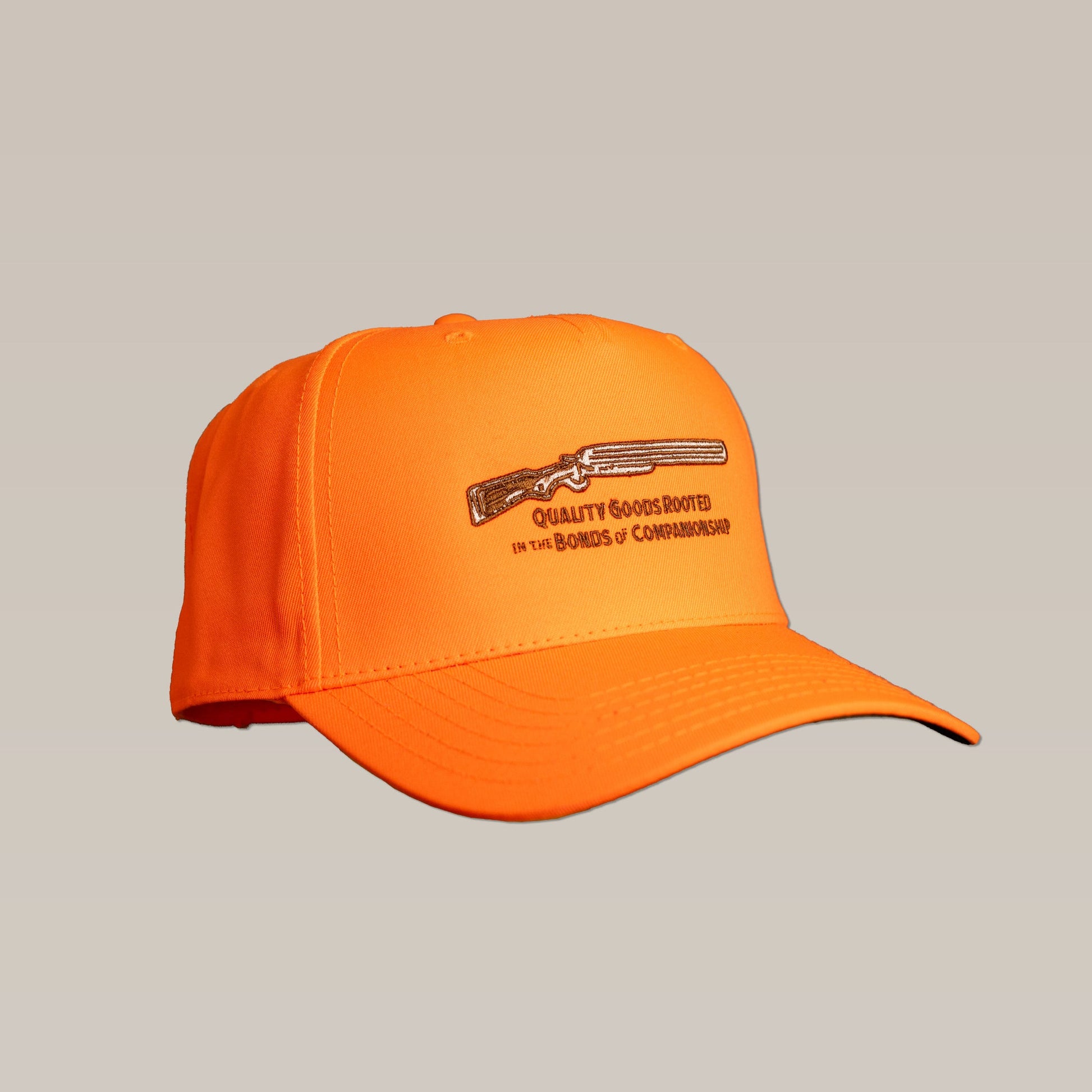 The Side by Side cap by Ballad of the Bird Dog is a bright orange baseball hat featuring an embroidered shotgun and the phrase "QUALITY GOODS ROOTED IN THE BONES OF COMPANIONSHIP" on the front.