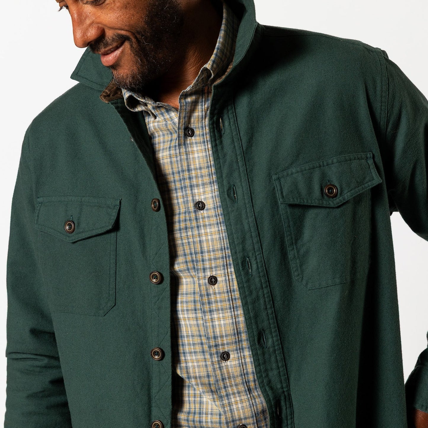 A man wears the Duck Head Slater Moleskin Overshirt as a sturdy outer layer over a plaid shirt, smiling and looking down against a white background.