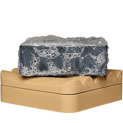 A gray, rectangular Dr.Squatch Soap Saver rests on a beige bricc soap dish with a carved pattern—an elegant accessory for any home.