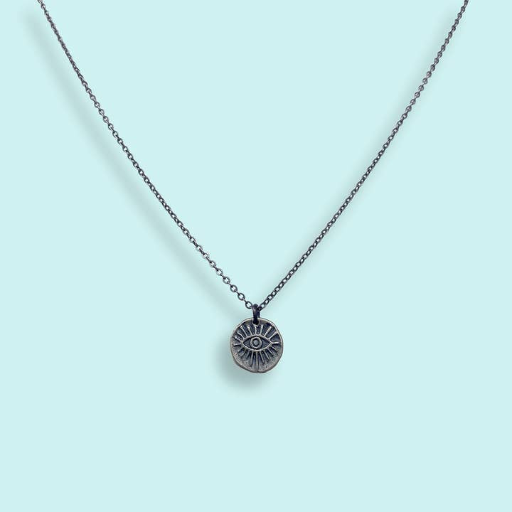 The Sterling Silver Evil Eye Medallion Necklace by Ornamental Things features a circular engraved eye pendant, shown against a light blue background.