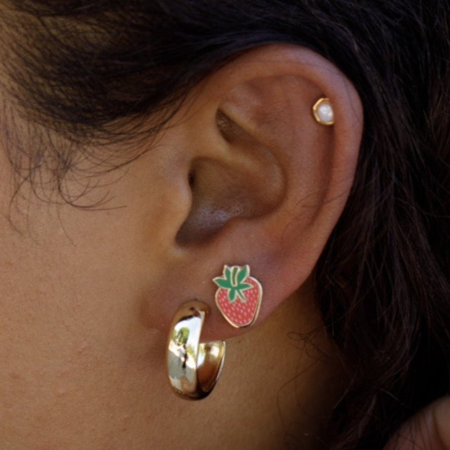 A close-up of an ear with three earrings: a small pearl stud in the cartilage, Larissa Loden’s Strawberry Enamel Studs, and a chunky gold hoop in the lobe.