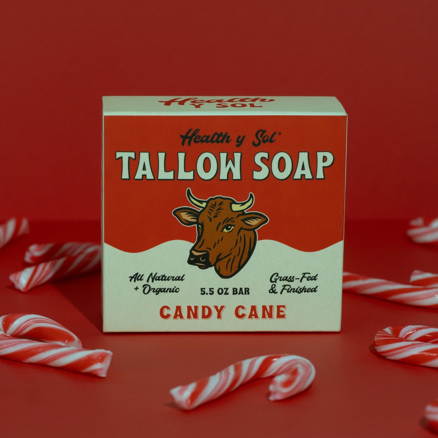 A box of Health y Sol Tallow Soap, made with all-natural ingredients and candy cane scent, sits on a red surface with candy canes. The red and white box features a cow illustration and helps reduce acne.