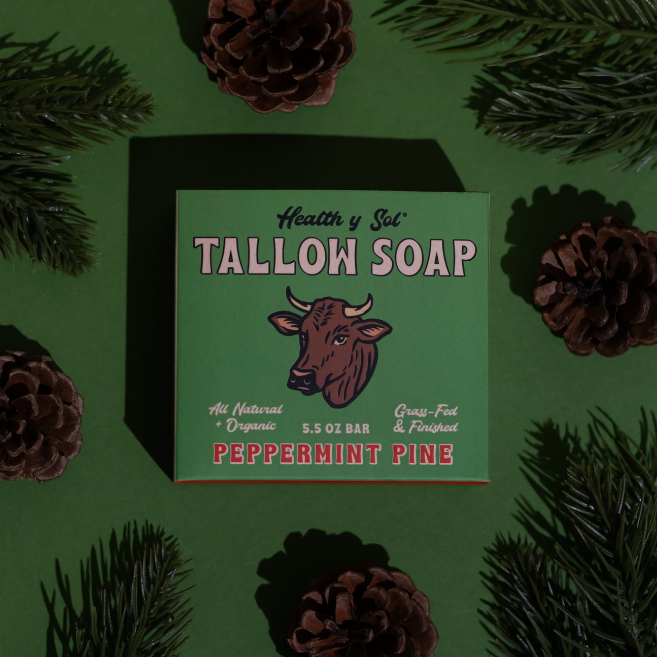 A green box of Health y Sol Tallow Soap in Peppermint Pine scent, made with all-natural ingredients, sits among pine branches and cones on a green background.