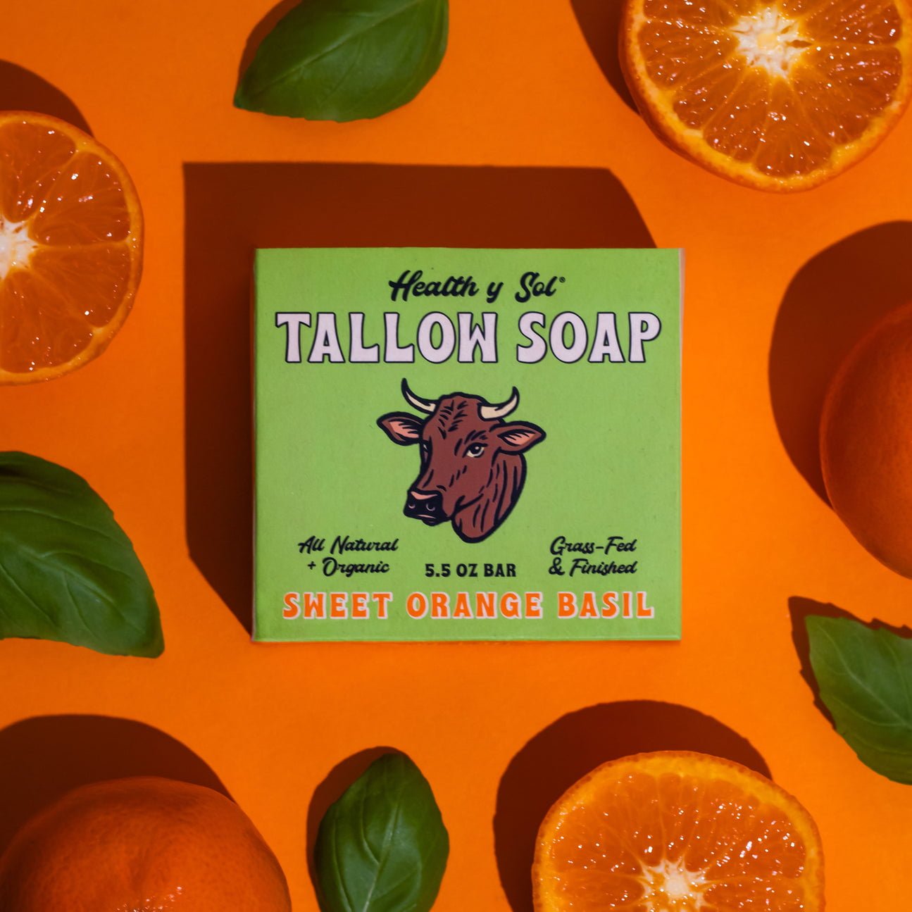 A box of Health y Sol Tallow Soap in sweet orange basil scent, made with natural ingredients, sits on an orange background surrounded by fresh basil leaves and halved oranges.
