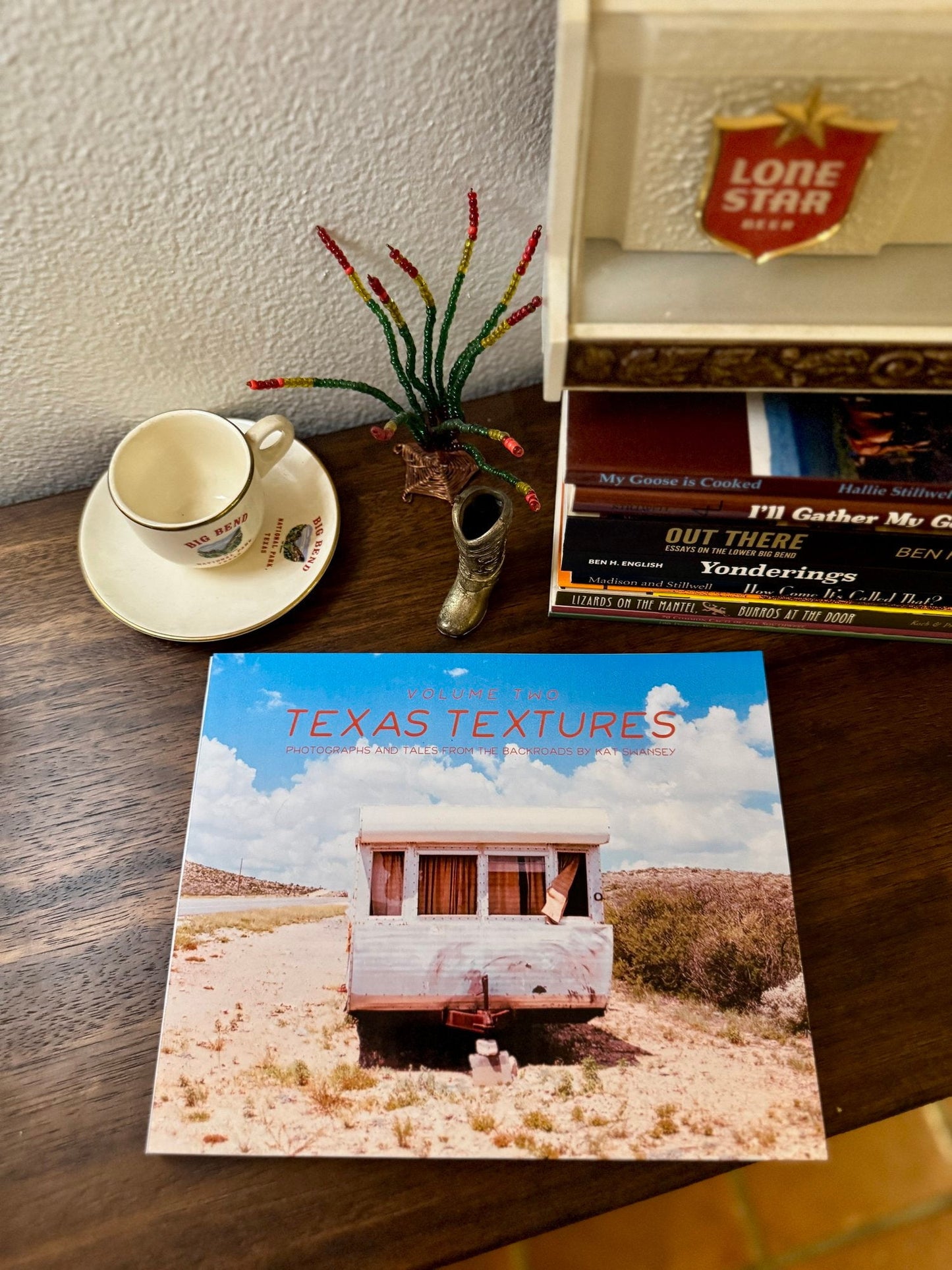 The "Texas Textures, Volume Two Book" by Kat Swansey rests on a wooden table next to a decorative cup, saucer, and beaded cactus ornament, featuring Texas photography with a trailer photo.