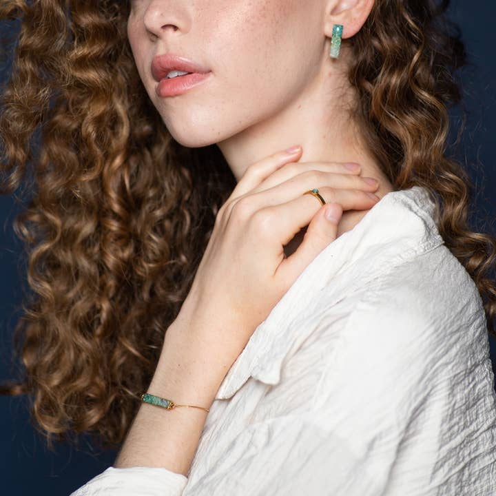 A woman with curly hair wears a white shirt and showcases green gemstone earrings, a ring, and the Colorblock Ombre Adjustable Bracelet by cameoko with her hand near her neck.
