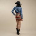 A person stands facing away in The Raquel Skirt by Sendero Provisions Co., styled with a black cowboy hat, blue shirt, and black cowboy boots against a neutral background.