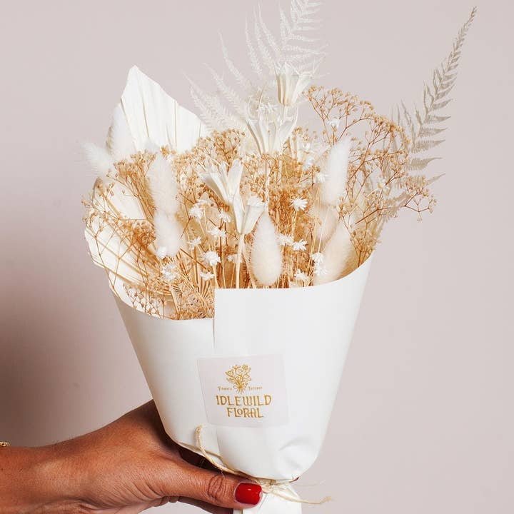 A hand holding The Snow Mini Preserved Bouquet by Idlewild Floral Co., a small arrangement of dried white and beige flowers, wrapped in white paper.