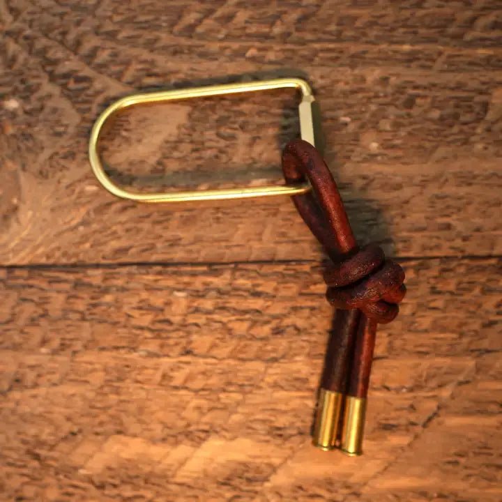 The Thistle Keychain by Tres Cuervos features a brass body with a brown leather loop and two brass-capped leather ends, shown resting on a wooden surface.