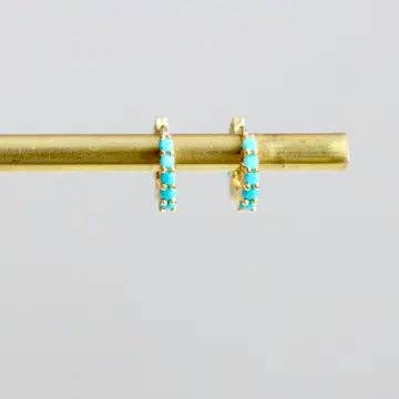 The Turquoise Huggie Earrings by Mesa Blue feature gold plating and turquoise stones, shown on a horizontal gold rod against a plain light background.