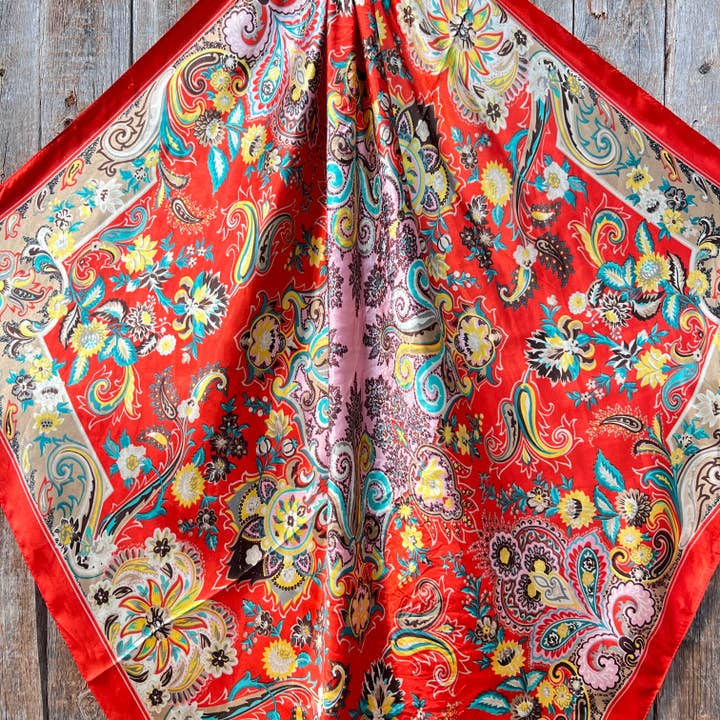 The Rodeo Drive "Turquoise That's So 70's Paisley Wild Rag" is a square silk scarf with vibrant floral and paisley designs on a turquoise background, displayed flat on a wooden surface.