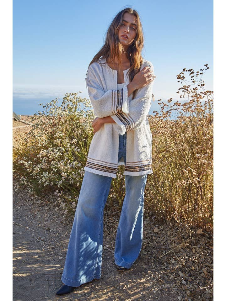 A woman stands outdoors among dry plants under a clear blue sky, wearing the By Together Tweed Long Sleeve Jacket paired with a white top and wide-leg blue jeans.