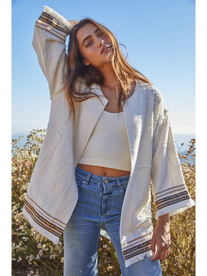 A woman stands outdoors with one arm raised against dry vegetation and a clear sky, wearing the By Together Tweed Long Sleeve Jacket paired with a ribbed white tank top and blue jeans.