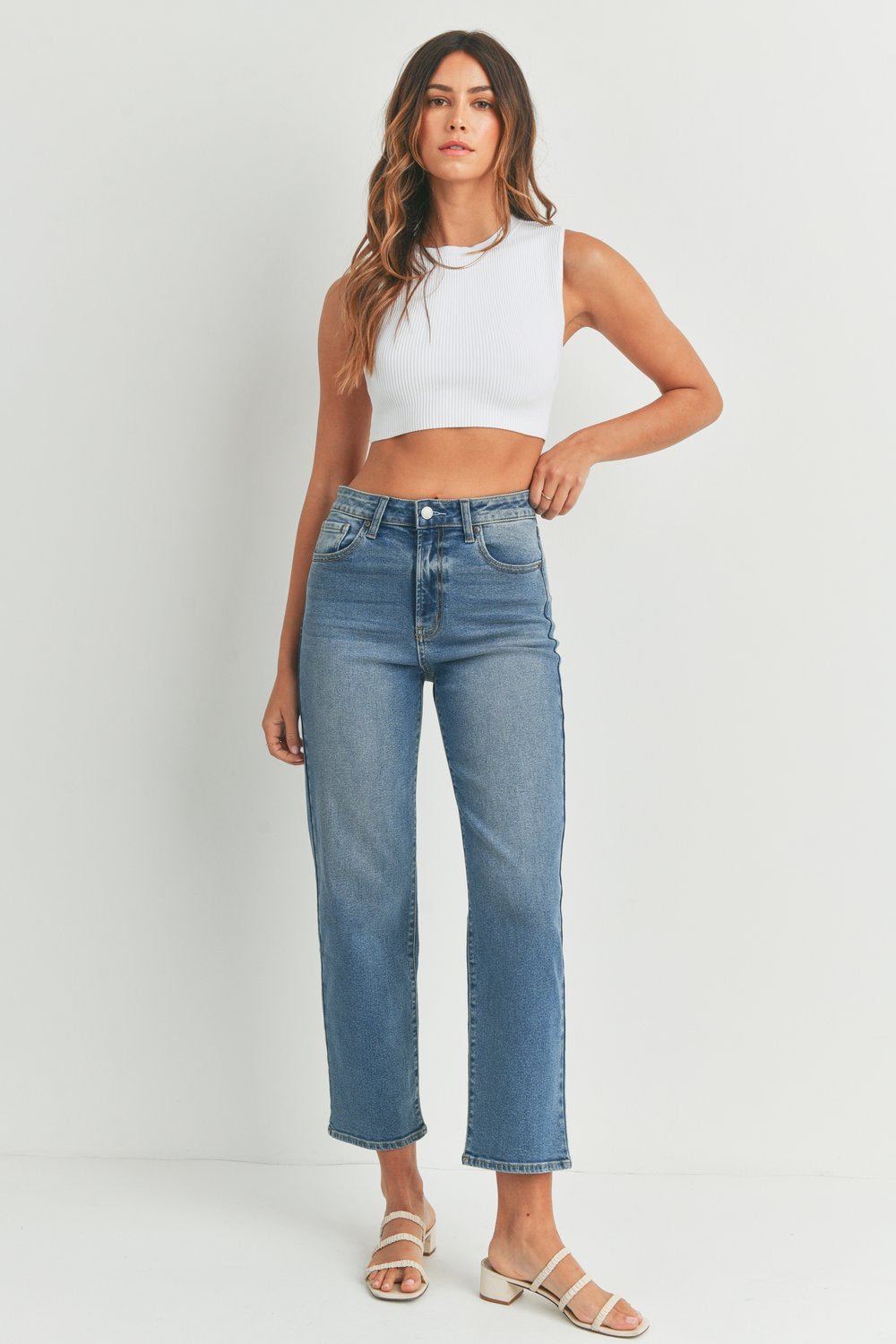 A woman stands against a white background wearing a white sleeveless crop top, Vintage Straight Jeans by JBD in medium denim, and white sandals.
