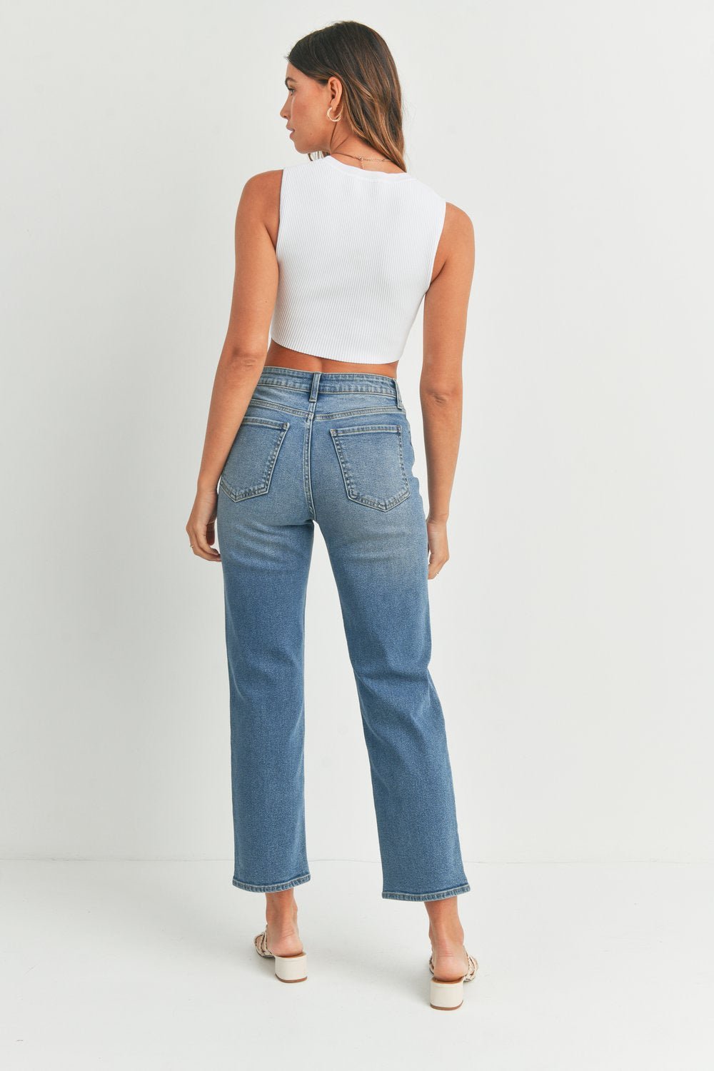 A woman faces away, wearing a white sleeveless crop top, JBD Vintage Straight Jeans in medium denim, and white shoes against a plain white background.