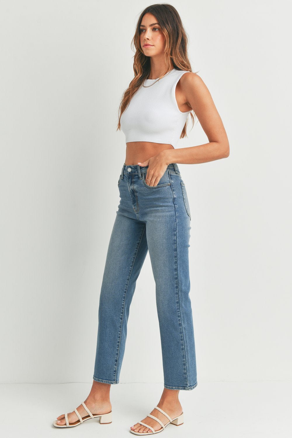 A woman stands against a white background, wearing a white sleeveless crop top, JBD Vintage Straight Jeans in medium denim, and white sandals.
