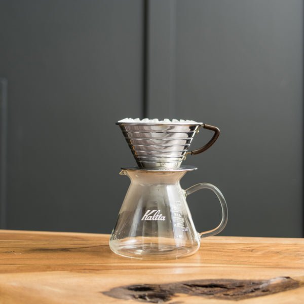 A Kalita pour-over dripper with a filter rests on the Kalita Wave Glass Coffee Server 500mL, made of heat-resistant glass with an ergonomic handle, placed on a wooden table against a plain dark background.