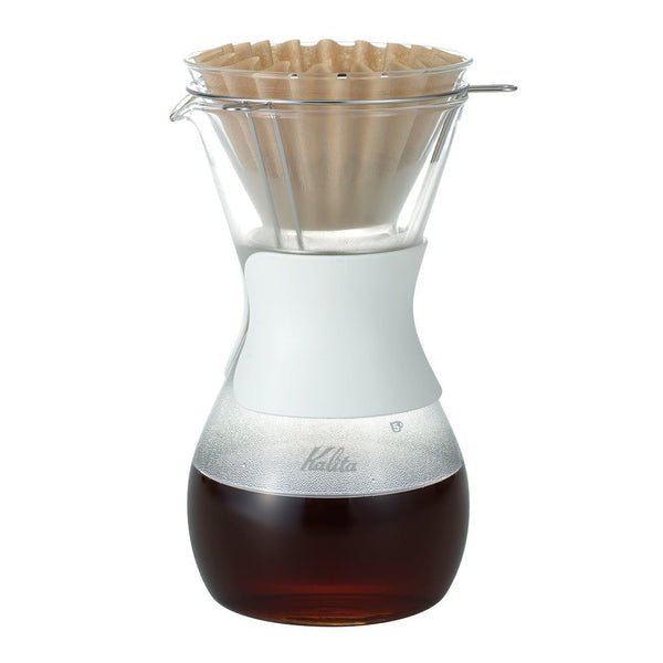 The Kalita Wave Style Glass Coffee Brewer, made from heat-resistant glass with a white silicone grip, is filled with brewed coffee and topped with a paper filter.