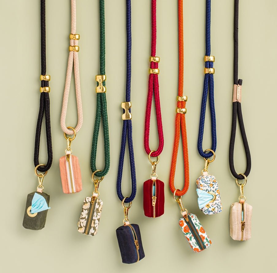 Eight colorful Waxed Canvas Poop Bag Dispensers by The Foggy Dog hang vertically, each featuring unique fabric patterns, gold hardware, and a zippered pouch design, displayed against a light green background.