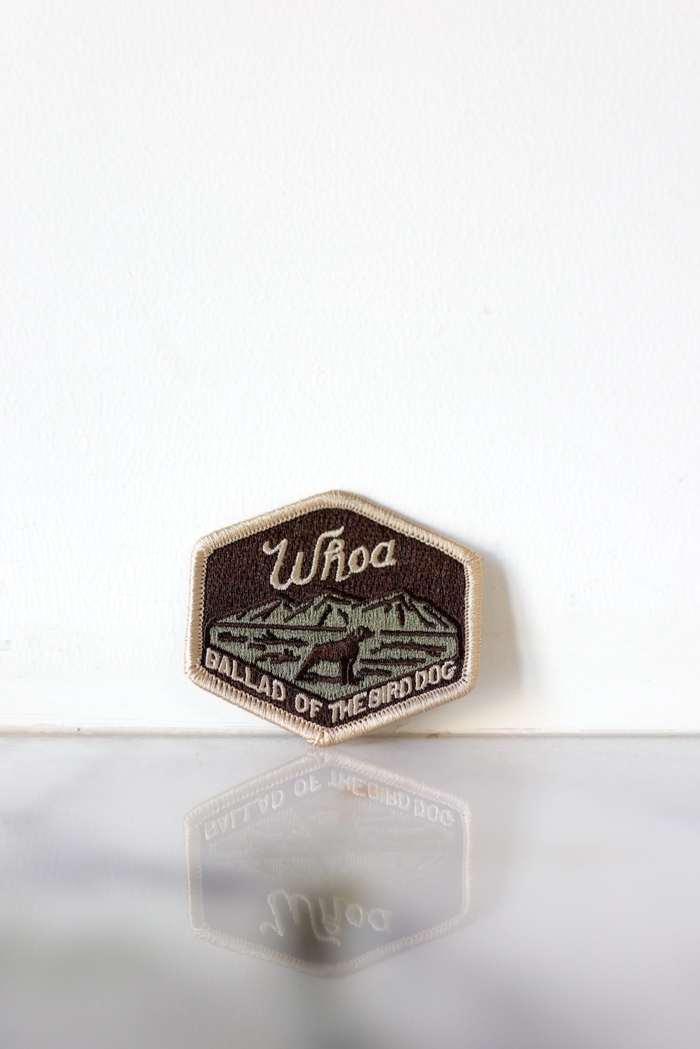 The Ballad of the Bird Dog Woah Patch, a hexagonal embroidered patch with "Woah" and a mountain design, stands upright against a white background.