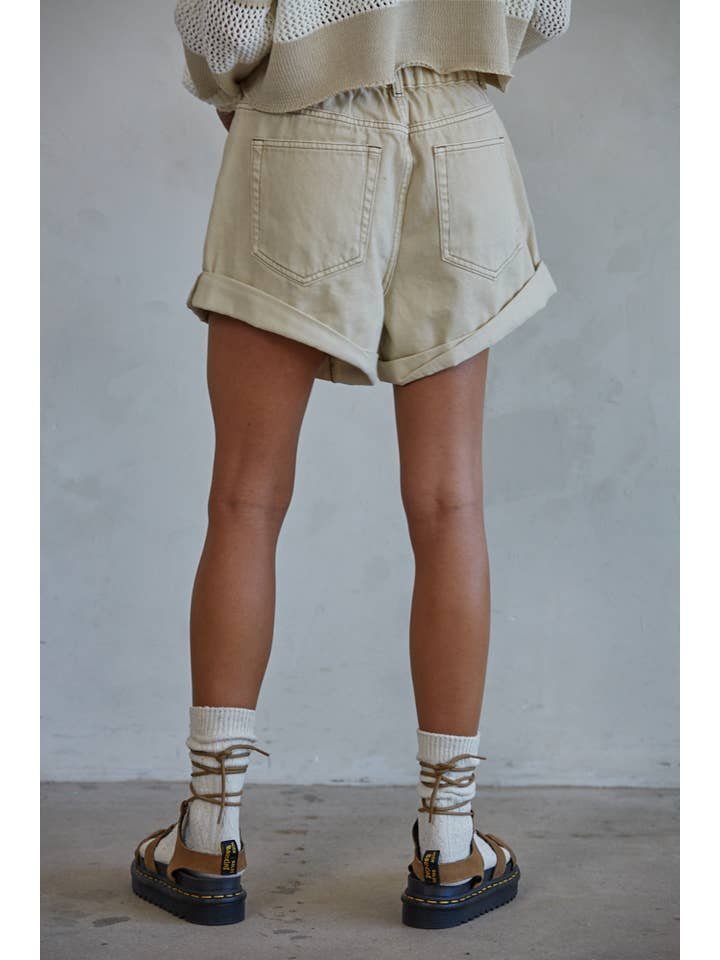 Person standing indoors, seen from behind, wears a loose-knit sweater, By Together Woven Cotton Denim Shorts in light beige rolled at the hem, white socks, and brown strapped sandals. The background is a plain light wall.
