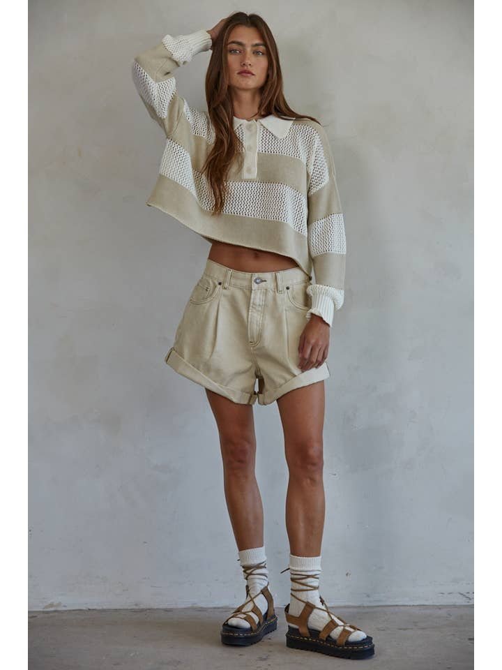 A woman stands against a plain wall wearing high-waisted Woven Cotton Denim Shorts by By Together, paired with a cropped, long-sleeve beige-and-white striped sweater, socks, and brown sandals.