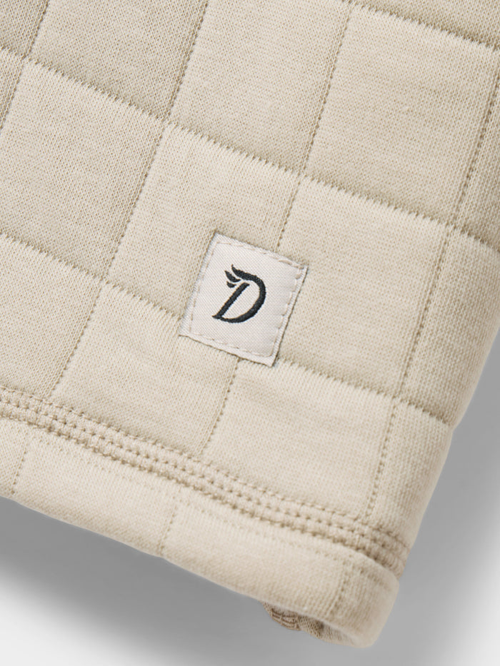 Close-up of the Duck Camp W's Cheyenne Quilted Crew, a beige cotton crew neck sweater featuring a stitched label with a stylized "D" and wing—ideal as a cozy layering piece.
