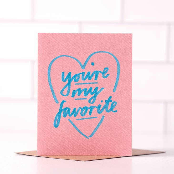 The "You're My Favorite" card by Daydream Prints features a pink blank interior, blue heart outline, and handwritten text on the front. It comes with a recycled envelope and is shown against a white tiled background.