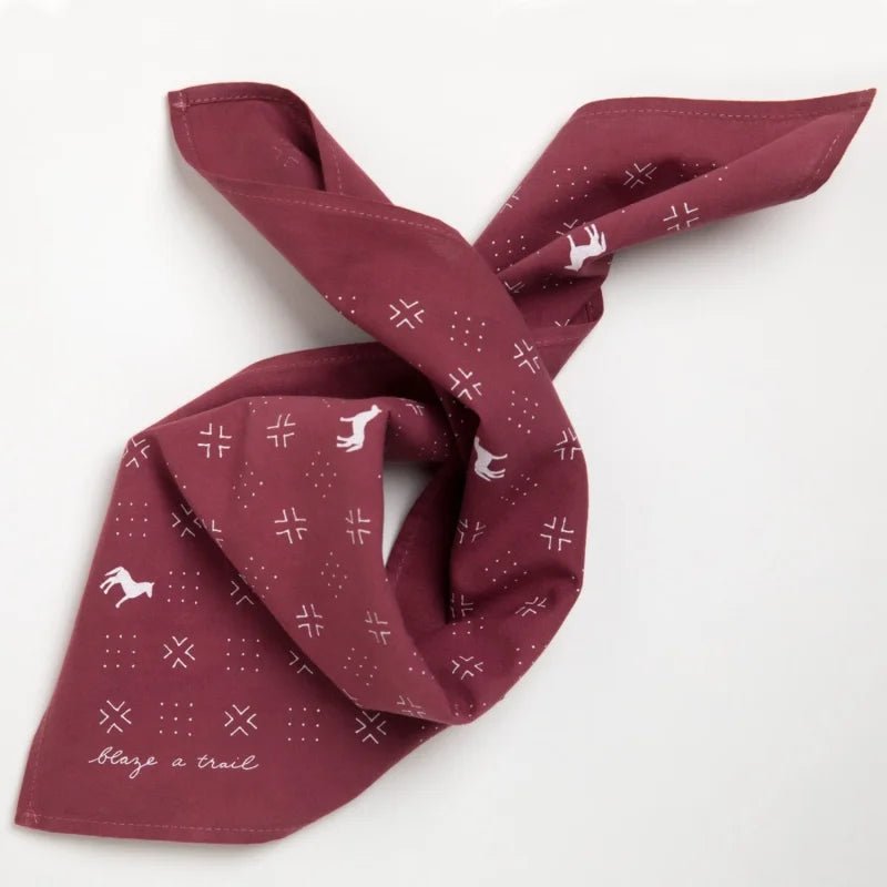 The Jenni Earle "Blaze A Trail" Bandana is hand-dyed maroon with white horse and cross patterns, partially folded, and features the phrase "blaze a trail" in the corner.