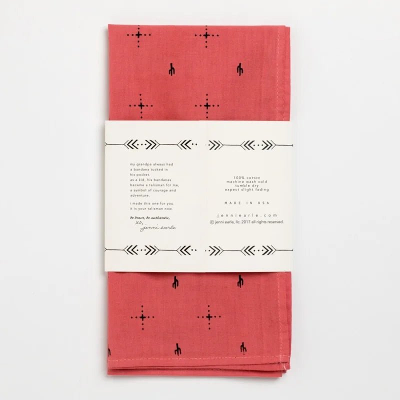 A folded red Jenni Earle "Explore More" cotton bandana with black geometric and horse designs, hand-dyed and wrapped in a white paper band with printed text.