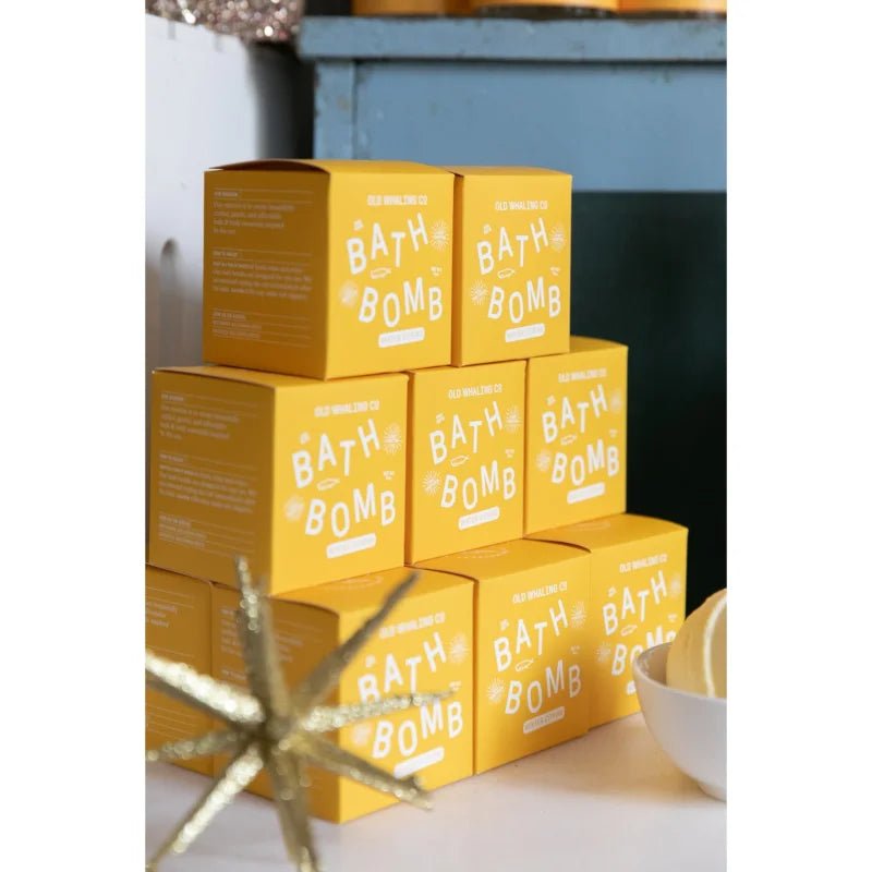 A stack of six Old Whaling Co. Bath Bomb | Seaside Citrine boxes is arranged on a surface, with a decorative object and a bowl containing epsom salts and bath bombs nearby.