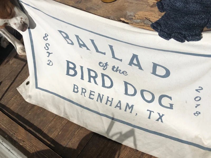 The Bird Dog Flag (Medium) by Ballad of the Bird Dog is a white shop flag featuring a blue stamp logo with "Ballad of the Bird Dog, Brenham, TX, Estd 2018," shown on wood with part of a dog’s head at the edge.