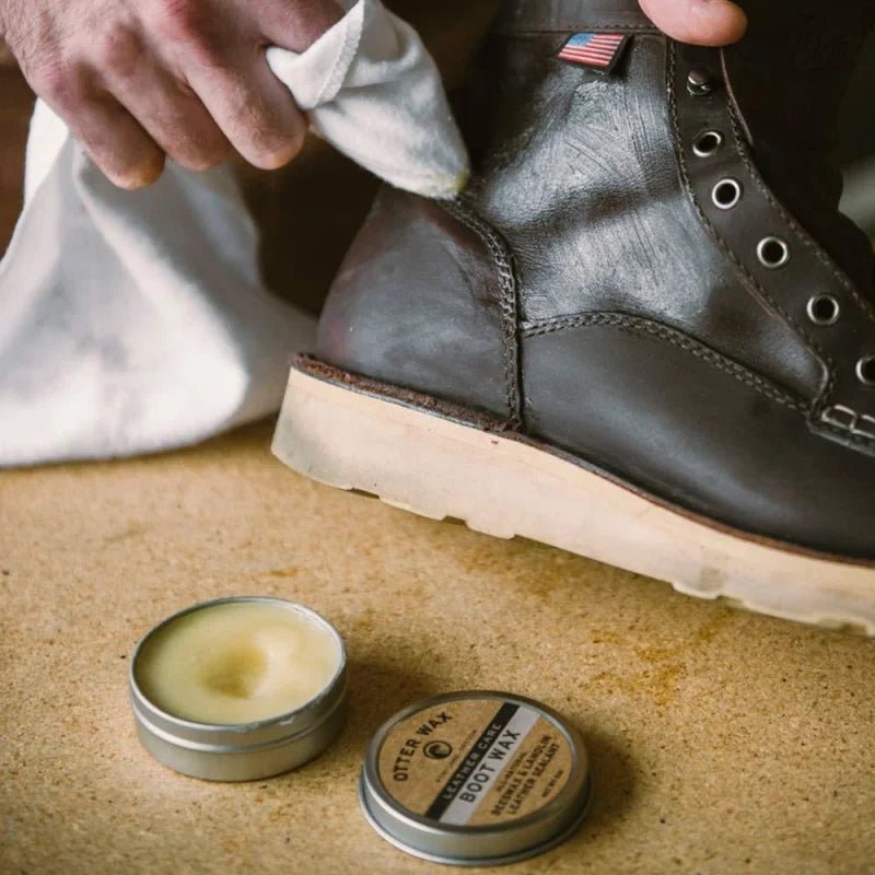 A person polishes a dark leather boot with a white cloth, while an open tin of Otter Wax Boot Wax by Otter Wax—famous for beeswax-based leather protection—rests nearby on a tan surface.