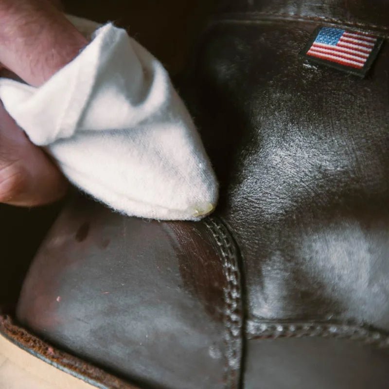 A hand uses a white cloth to polish a dark leather boot with an American flag patch, applying Otter Wax Boot Wax for added leather protection.