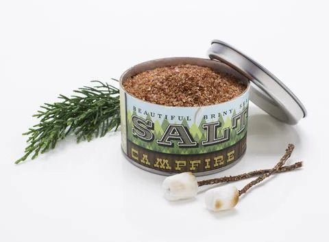 An open tin of Campfire Sea Salt by Beautiful Briny Sea, filled with smoked sea salt, sits beside greenery and two toasted marshmallows on sticks.