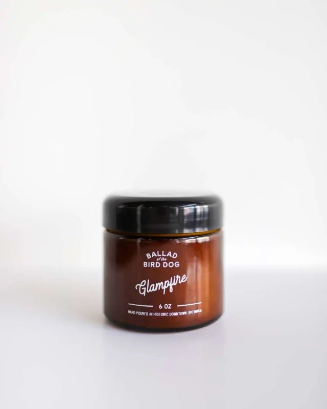 A brown glass jar candle with a black lid, labeled "Candle | Glampfire | Ballad of the Bird Dog" by Ballad of the Bird Dog, rests on a white surface, evoking Texas campfire vibes with its inviting Arabian wood scent.