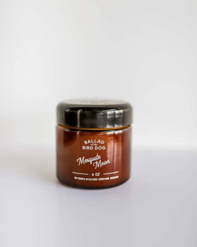A brown glass jar labeled "Candle | Mesquite Moon | Ballad of the Bird Dog" with a black lid rests on a white surface, capturing the warm, smoky scent of saffron tobacco by Ballad of the Bird Dog.