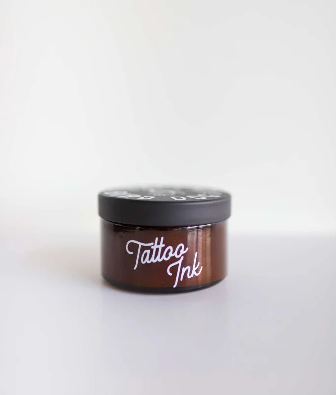 A small brown jar with a black lid labeled "Tattoo Ink" by Ballad of the Bird Dog sits against a white background, evoking a candle scent with notes of patchouli, vanilla, and musk.